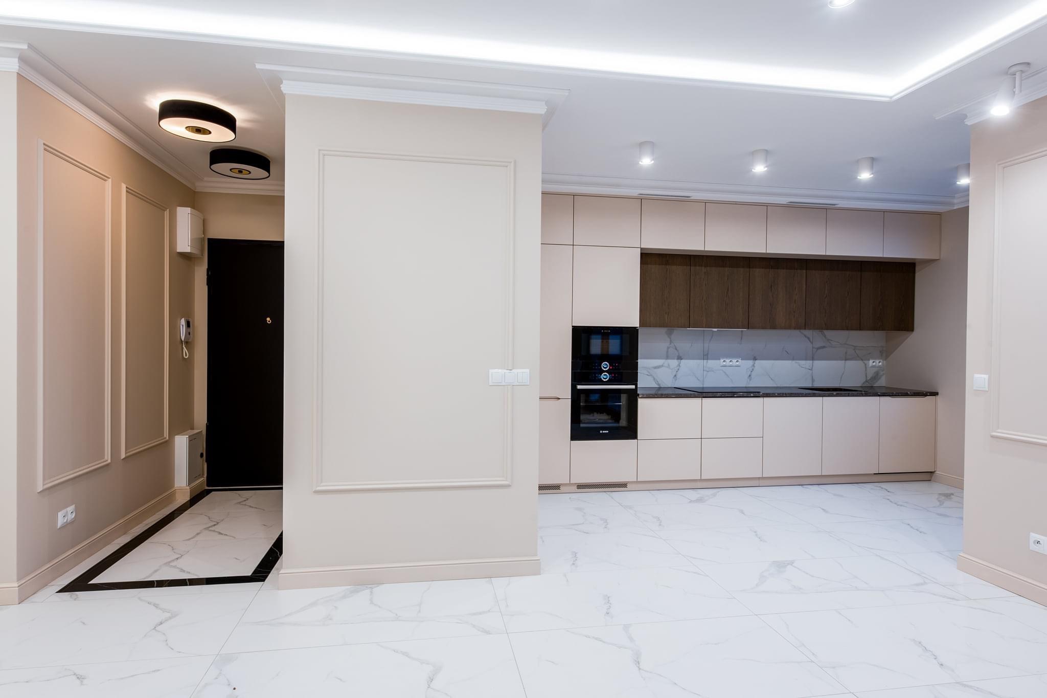 Nowoczesna kuchnia z jasnymi szafkami, ciemnymi akcentami drewna i marmurową podłogą, widok na przedpokój z czarnymi elementami wykończenia.