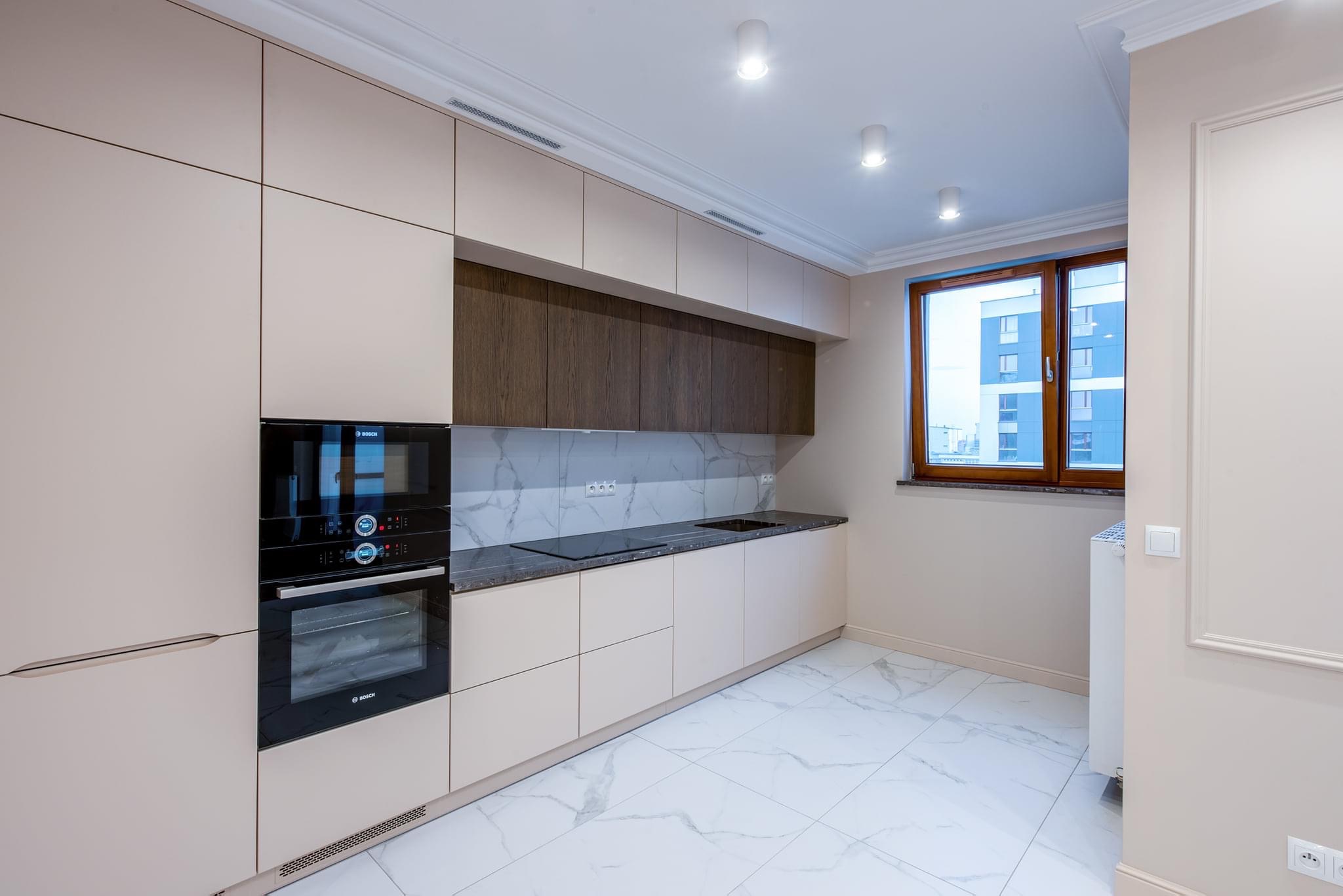 Nowoczesna kuchnia z beżowymi szafkami, marmurowym blatem i białymi płytkami podłogowymi w jodełkę, widok na budynek z okna.