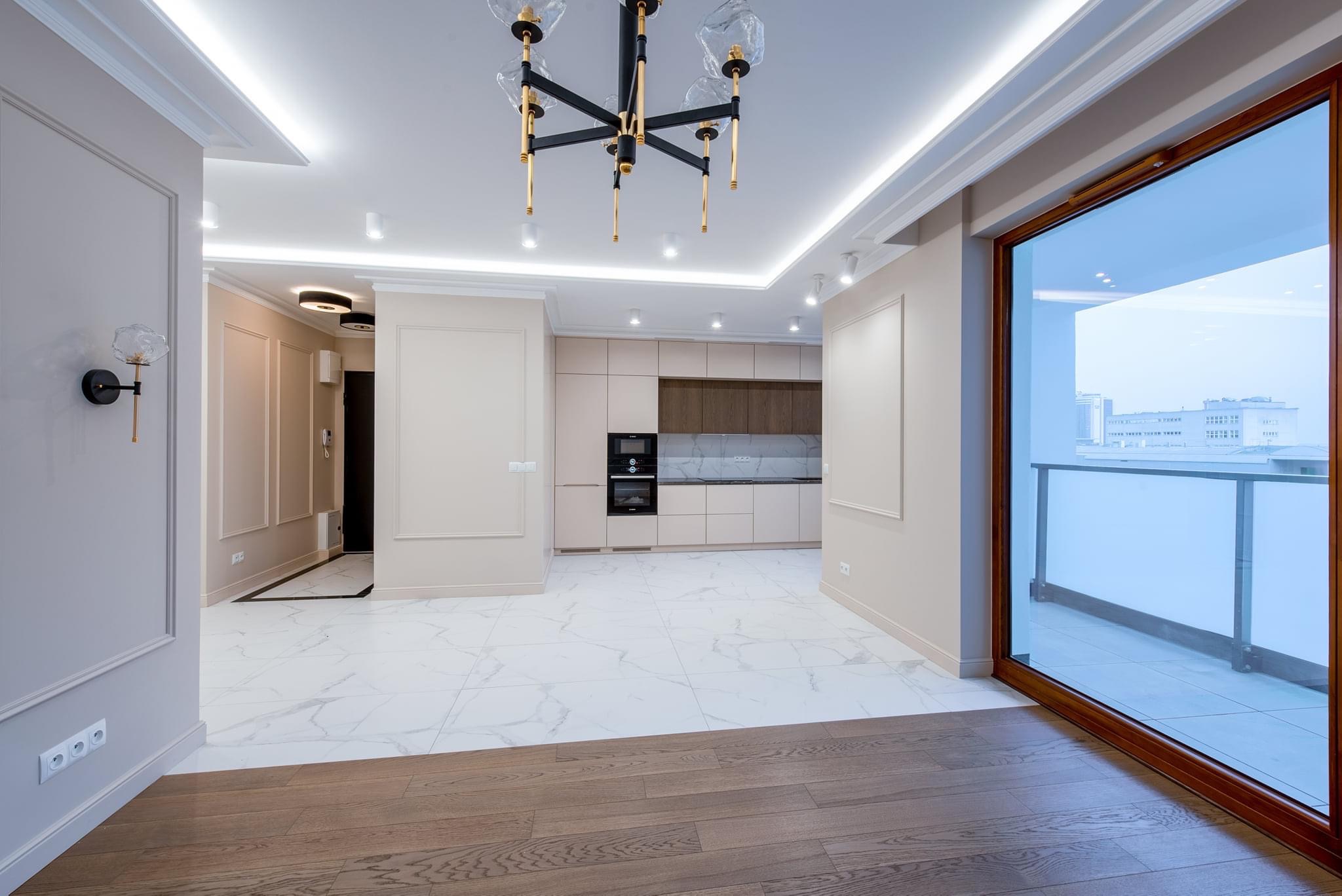 Nowoczesne wnętrze po remoncie: połączenie drewnianej podłogi, białych marmurowych płytek, beżowych ścian z dekoracyjnymi listwami i otwartej kuchni z ciemnymi akcentami, widok na miasto przez duże...