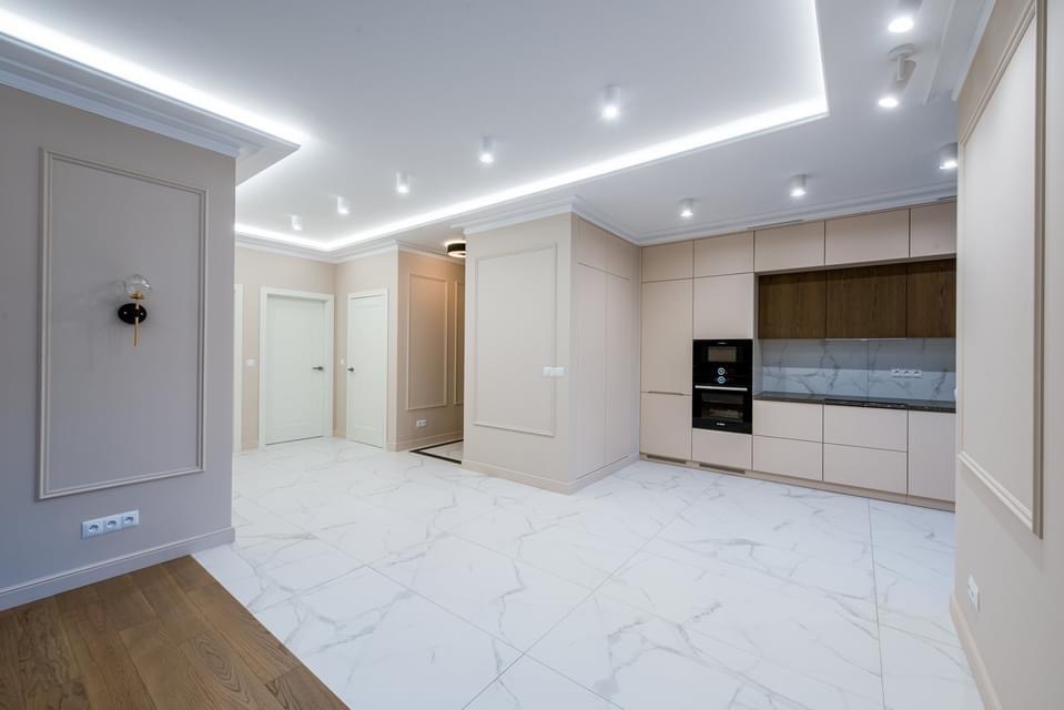 Przestronne wnętrze po remoncie: białe marmurowe płytki podłogowe, beżowe ściany z dekoracyjnymi listwami, zabudowa kuchenna w odcieniach beżu i brązu, oświetlenie sufitowe punktowe i ledowe.