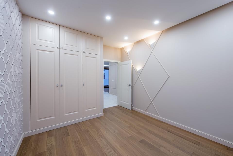 Jasne wnętrze pokoju z drewnianą podłogą, zabudowaną szafą w kolorze ecru, ścianą z dekoracyjną tapetą i ścianą z geometrycznym wzorem z listew.