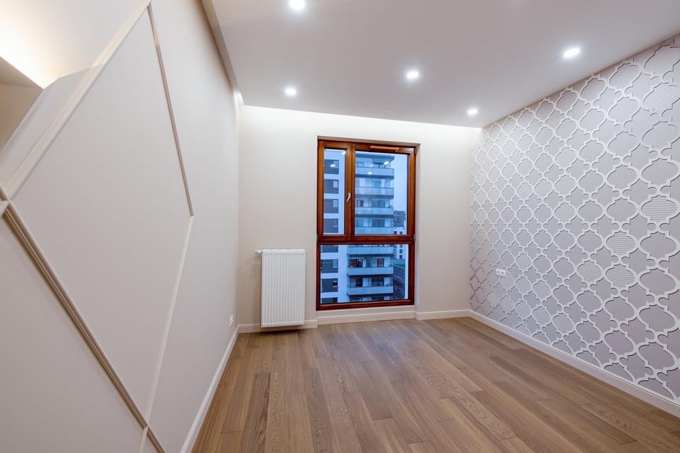 Jasne, puste pomieszczenie z drewnianą podłogą, wzorzystą tapetą w odcieniach szarości, białym grzejnikiem i oknem z widokiem na budynek mieszkalny.