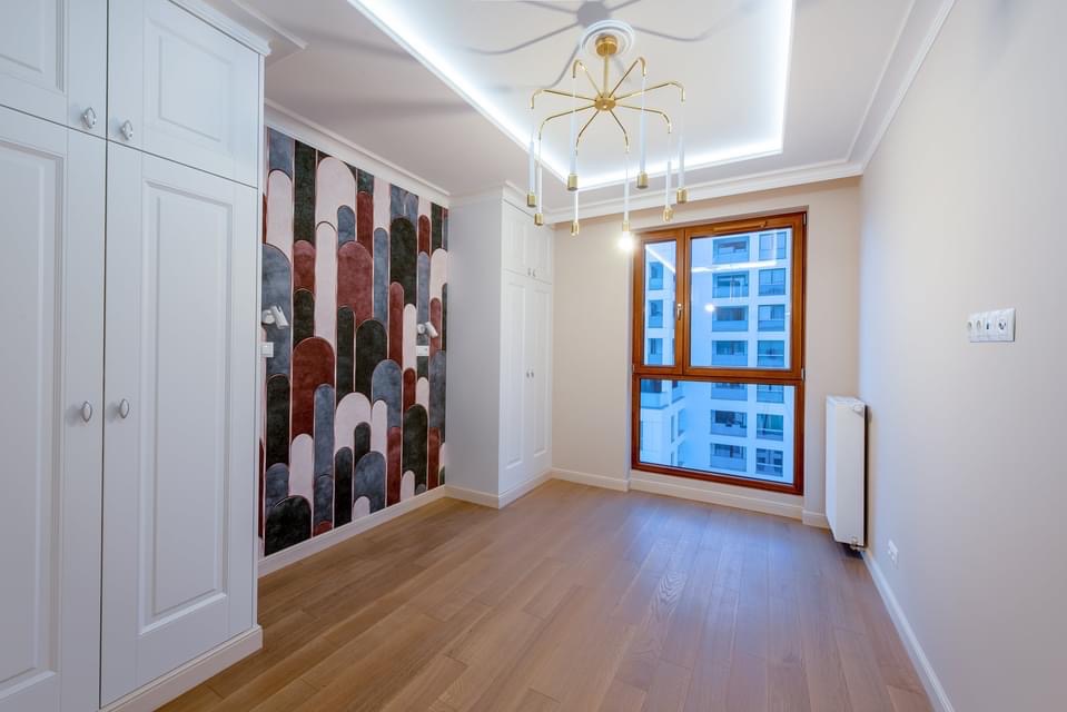 Wykończone wnętrze pokoju z parkietem, tapetą w geometryczne wzory, białymi szafami wnękowymi i nowoczesnym żyrandolem z mlecznymi kloszami.