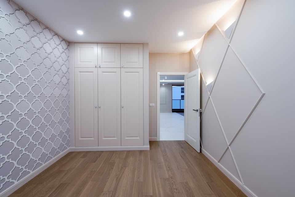 Jasne wnętrze pokoju po remoncie z drewnianą podłogą, szafą w zabudowie, ścianą z dekoracyjną tapetą w marokański wzór i ścianą z geometrycznymi listwami podświetlonymi od góry.