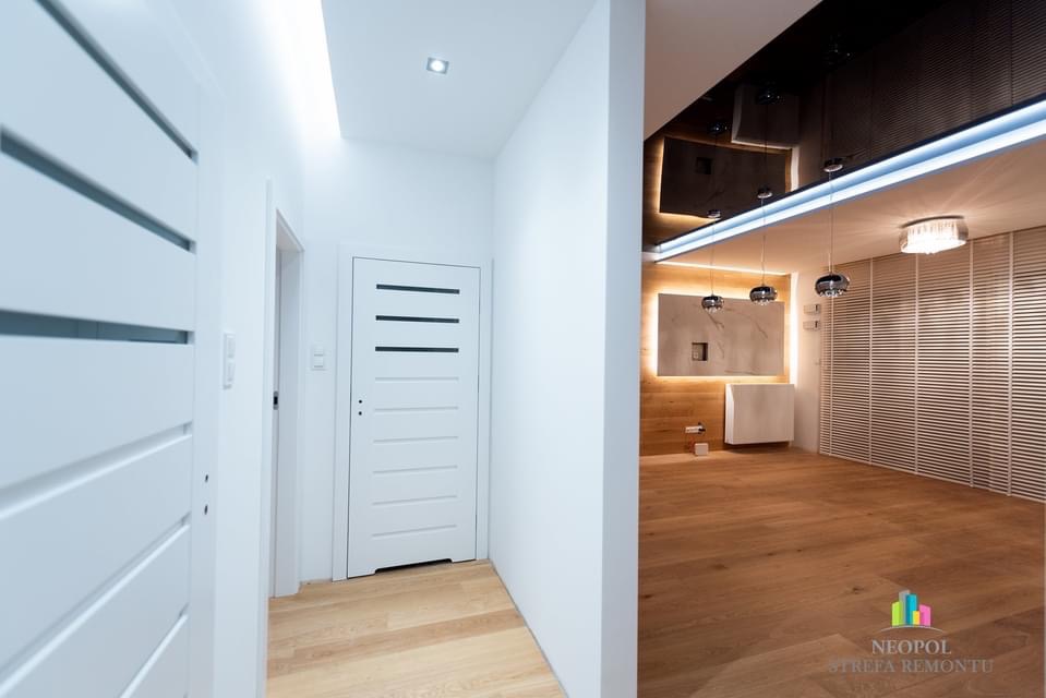 Nowoczesny korytarz z białymi drzwiami z poziomymi przeszkleniami i drewnianą podłogą, widok na łazienkę z lustrem podświetlonym od tyłu i drewnianymi panelami ściennymi.