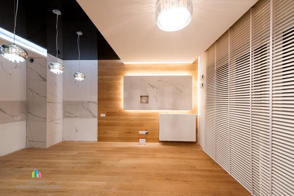 Nowoczesne wnętrze po remoncie: parkiet z jasnego drewna, ściany wykończone płytami imitującymi marmur, drewniana boazeria z podświetleniem LED wokół marmurowej płyty, białe żaluzje pionowe...