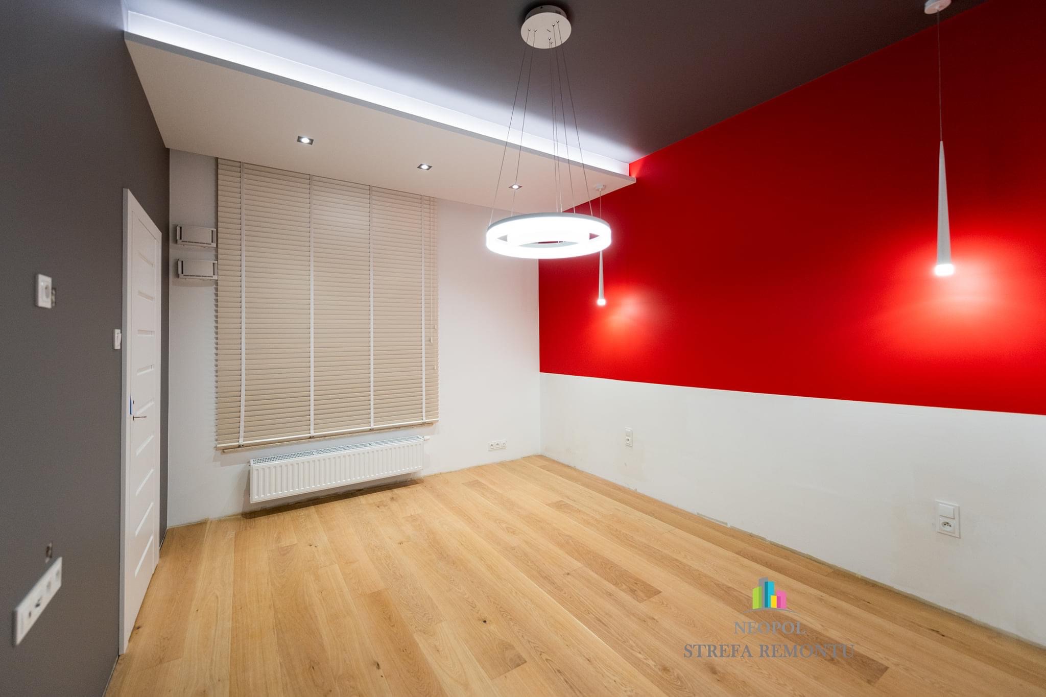 Wnętrze pokoju po remoncie: czerwona ściana kontrastująca z szarą, drewniana podłoga, nowoczesne oświetlenie z okrągłą lampą wiszącą i żaluzjami w oknie.