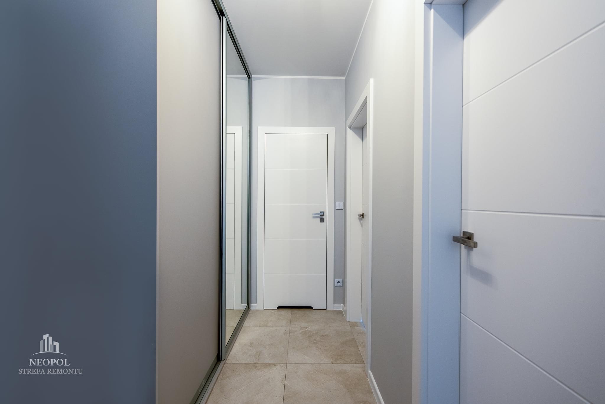 Nowoczesny korytarz z szarą szafą przesuwną z lustrem, białymi drzwiami wewnętrznymi z poziomymi frezami i beżową podłogą z płytek.