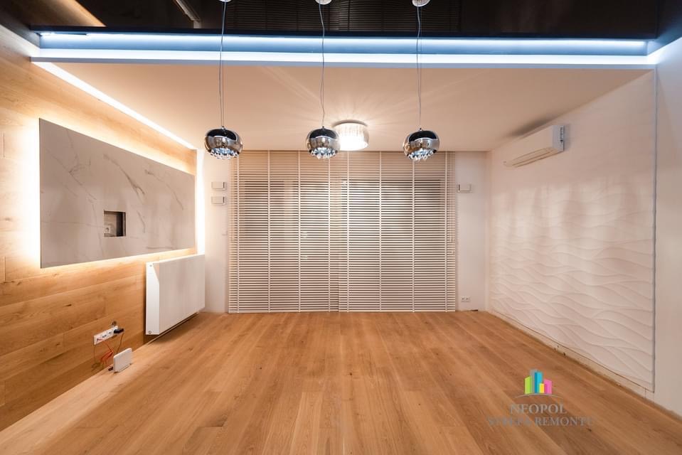 Jasne, przestronne wnętrze po remoncie z drewnianą podłogą, ścianą z paneli imitujących drewno i marmur z podświetleniem LED, żaluzjami poziomymi oraz dekoracyjną ścianą z motywem fal. Widoczne...