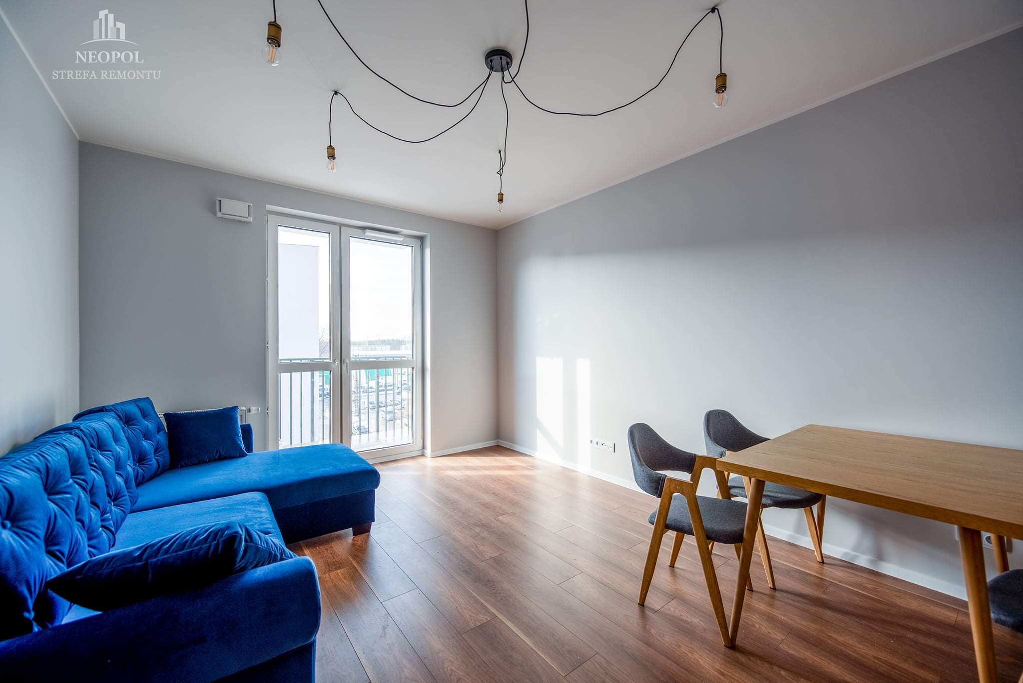 Jasne wnętrze po remoncie z parkietem, szarą ścianą, niebieską sofą i stołem z krzesłami, oświetlone lampami na kablach.