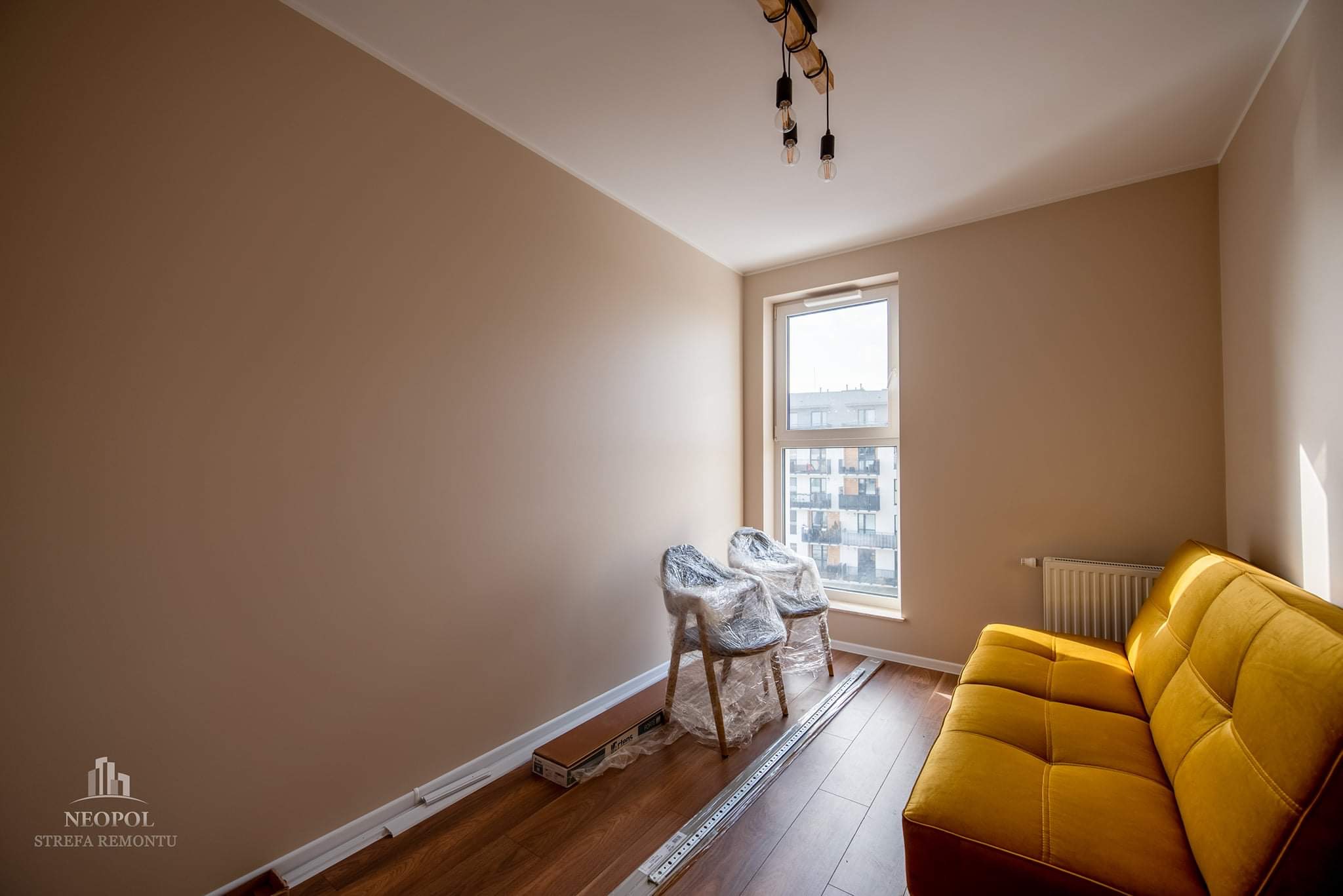 Pokój po malowaniu ścian na beżowo, z nową podłogą, widoczne dwa krzesła owinięte folią ochronną, żółta sofa i elementy do montażu listew przypodłogowych.