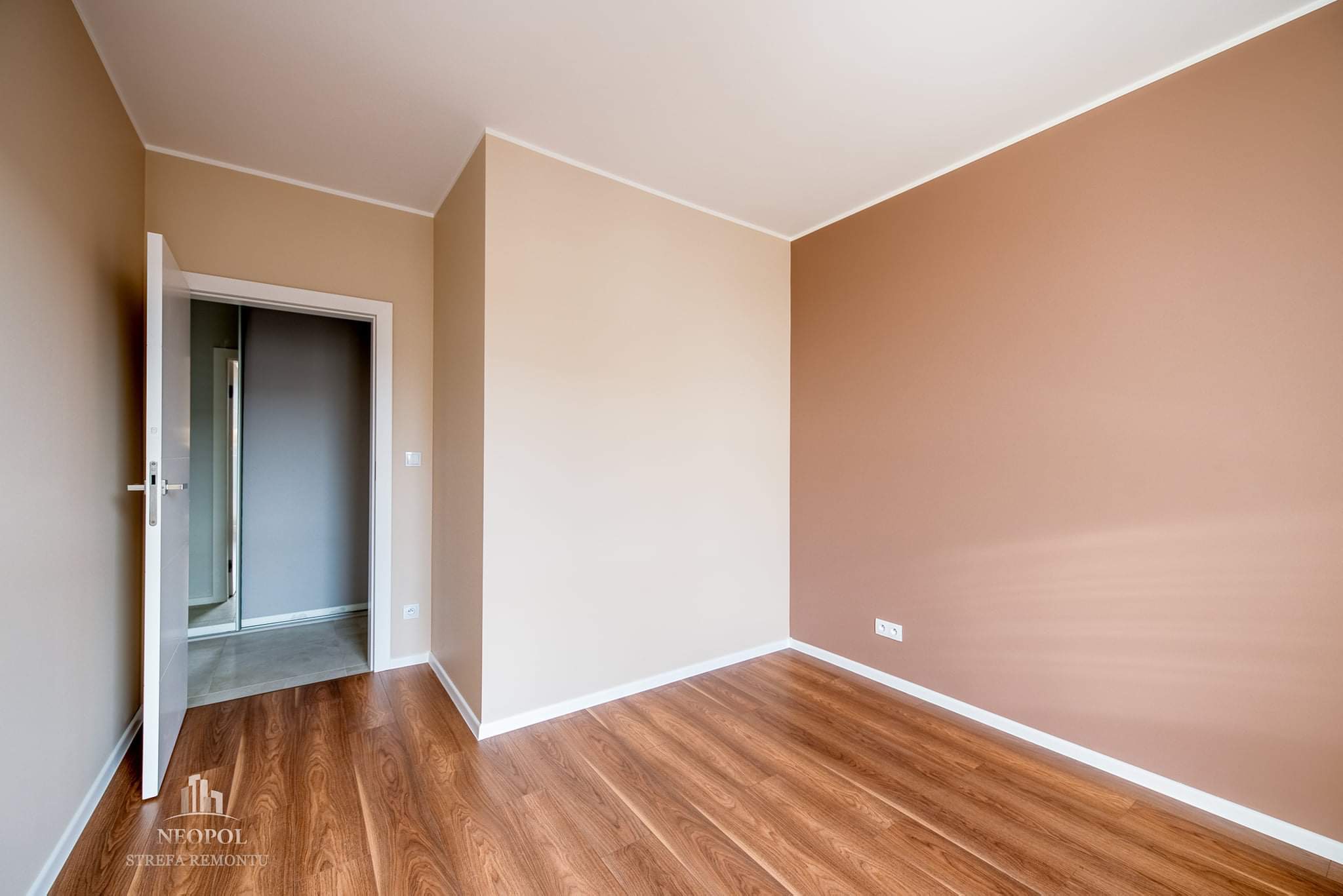 Pomalowane na dwa kolory wnętrze pokoju z drewnianą podłogą i otwartymi drzwiami, widoczne gniazdka elektryczne, listwy przypodłogowe. W rogu logo firmy.