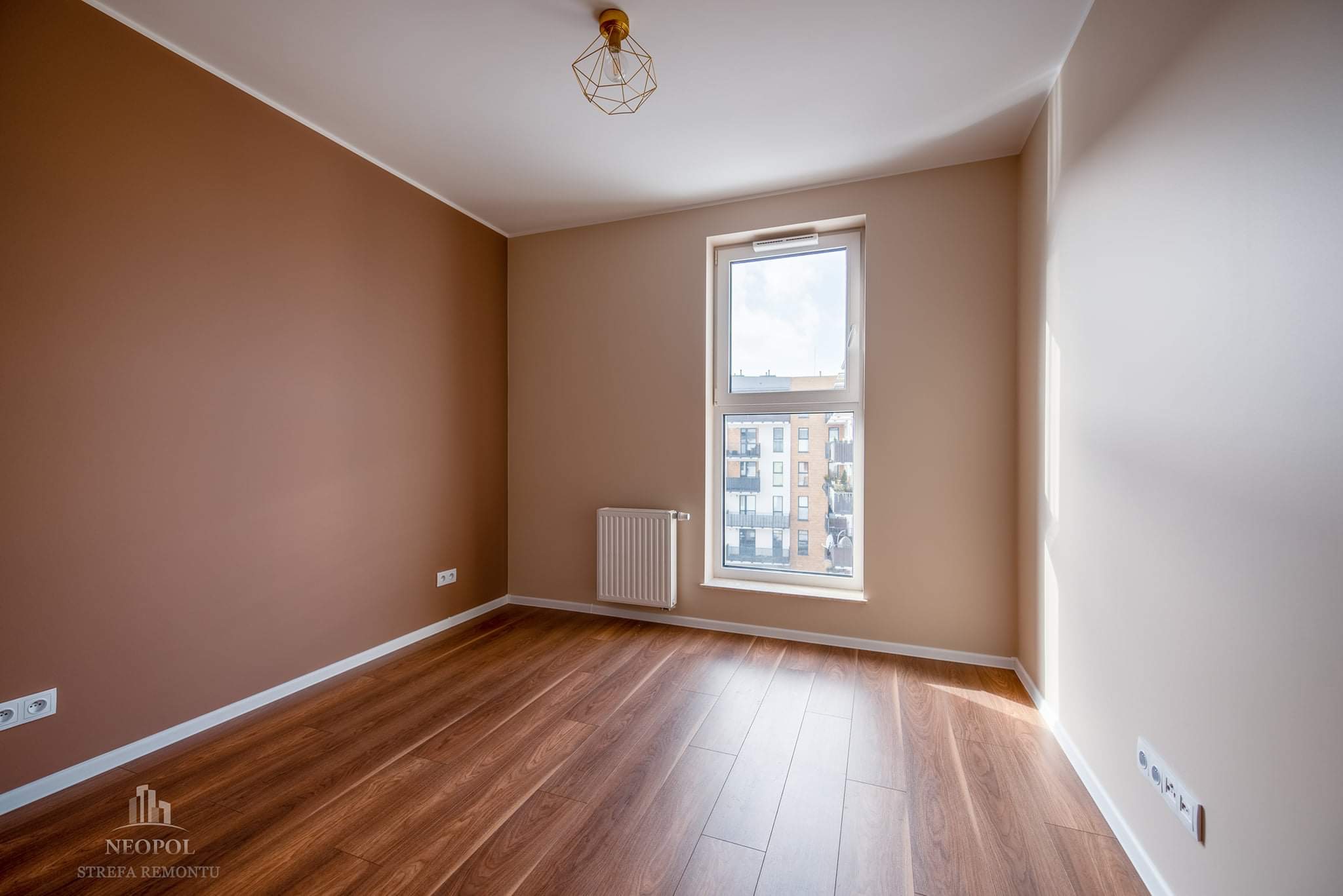 Pomalowane na brązowo i beżowo wnętrze pokoju z drewnianą podłogą, grzejnikiem pod oknem i nowoczesną lampą sufitową. Widoczne gniazdka elektryczne przy podłodze.
