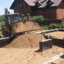 Żółta koparko-ładowarka z pełną łyżką piasku na tle fundamentów domu w trakcie budowy, widoczny człowiek w czerwonych spodenkach i biała butelka z płynem.