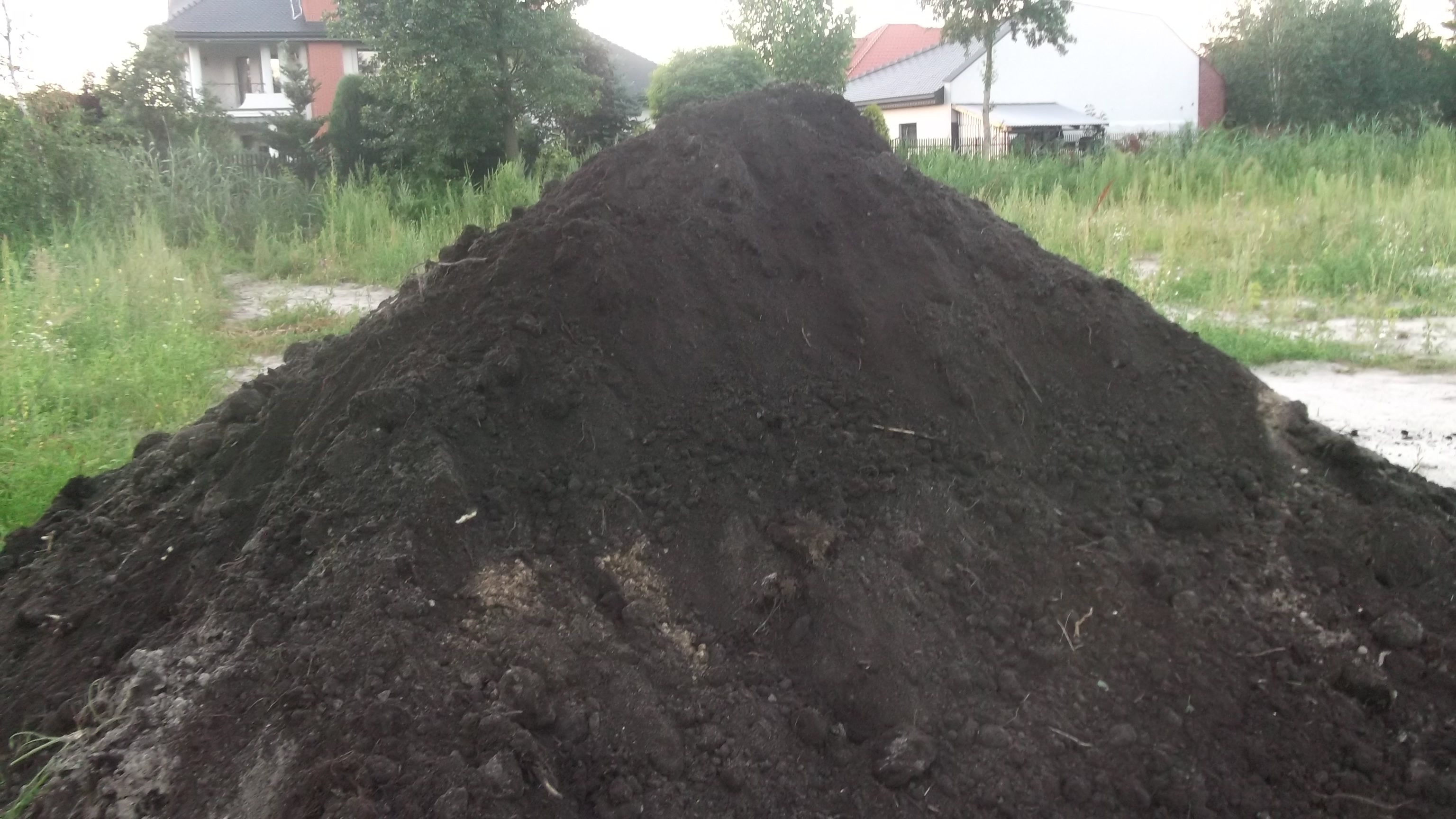 Kupa ciemnej ziemi na działce z widocznymi budynkami mieszkalnymi w tle.