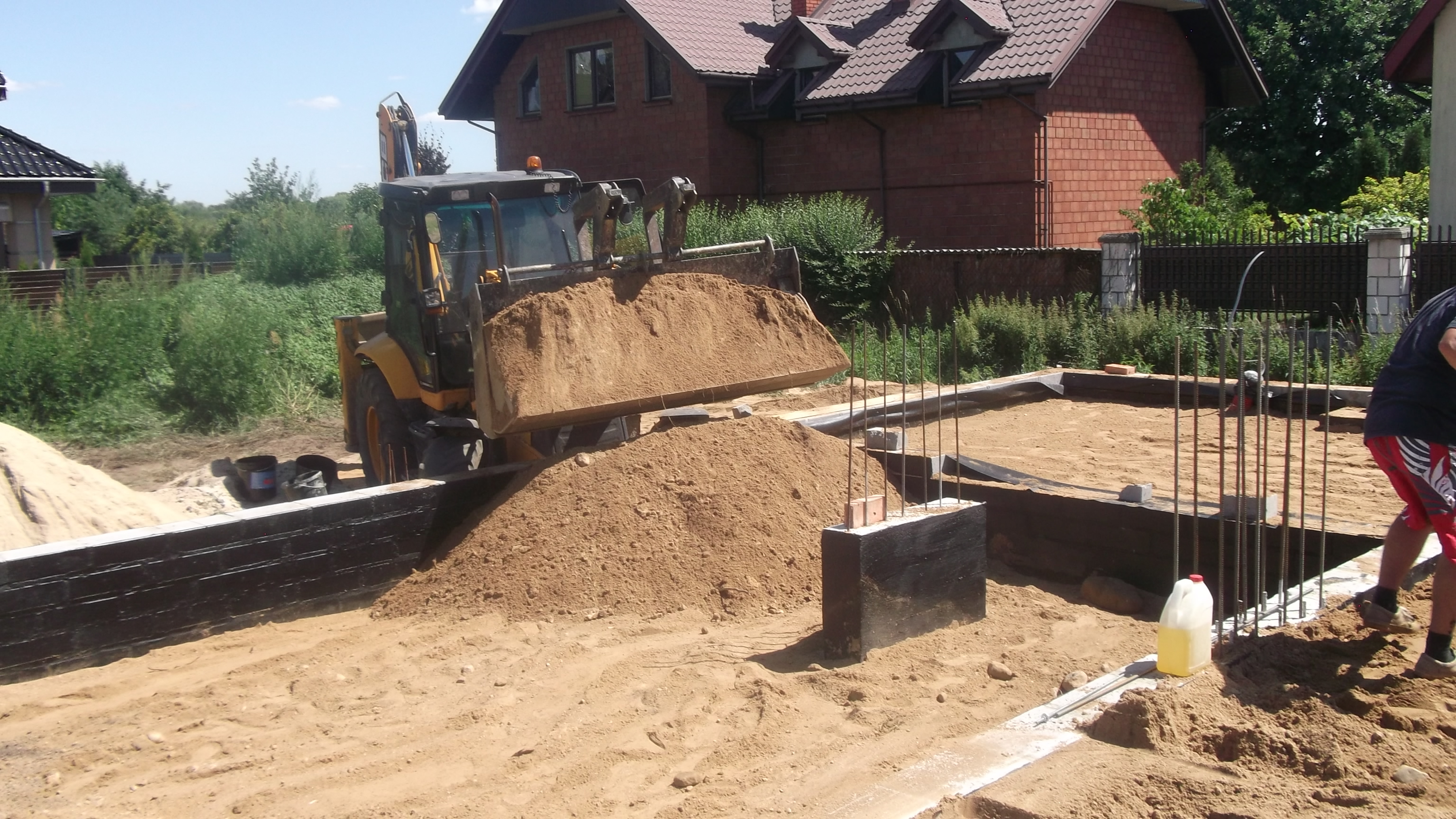 Żółta koparko-ładowarka z pełną łyżką piasku na tle fundamentów domu w trakcie budowy, widoczny człowiek w czerwonych spodenkach i biała butelka z płynem.