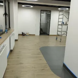 Wnętrze biura po remoncie: widoczne wiaderka z farbą, aluminiowa drabina na k&oacute;łkach, zabezpieczone taśmą malarską ściany, podłoga wyłożona płytkami imitującymi drewno.