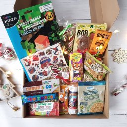 Firma Usługowa Aleksandra Janusz - Otwarty karton wypełniony różnorodnymi słodyczami, przekąskami i zabawkami dla dzieci, w tym lizaki, ciasteczka kokosowe, batony owocowe, książeczka do zdrapywania z motywem Minecrafta i naklejki...