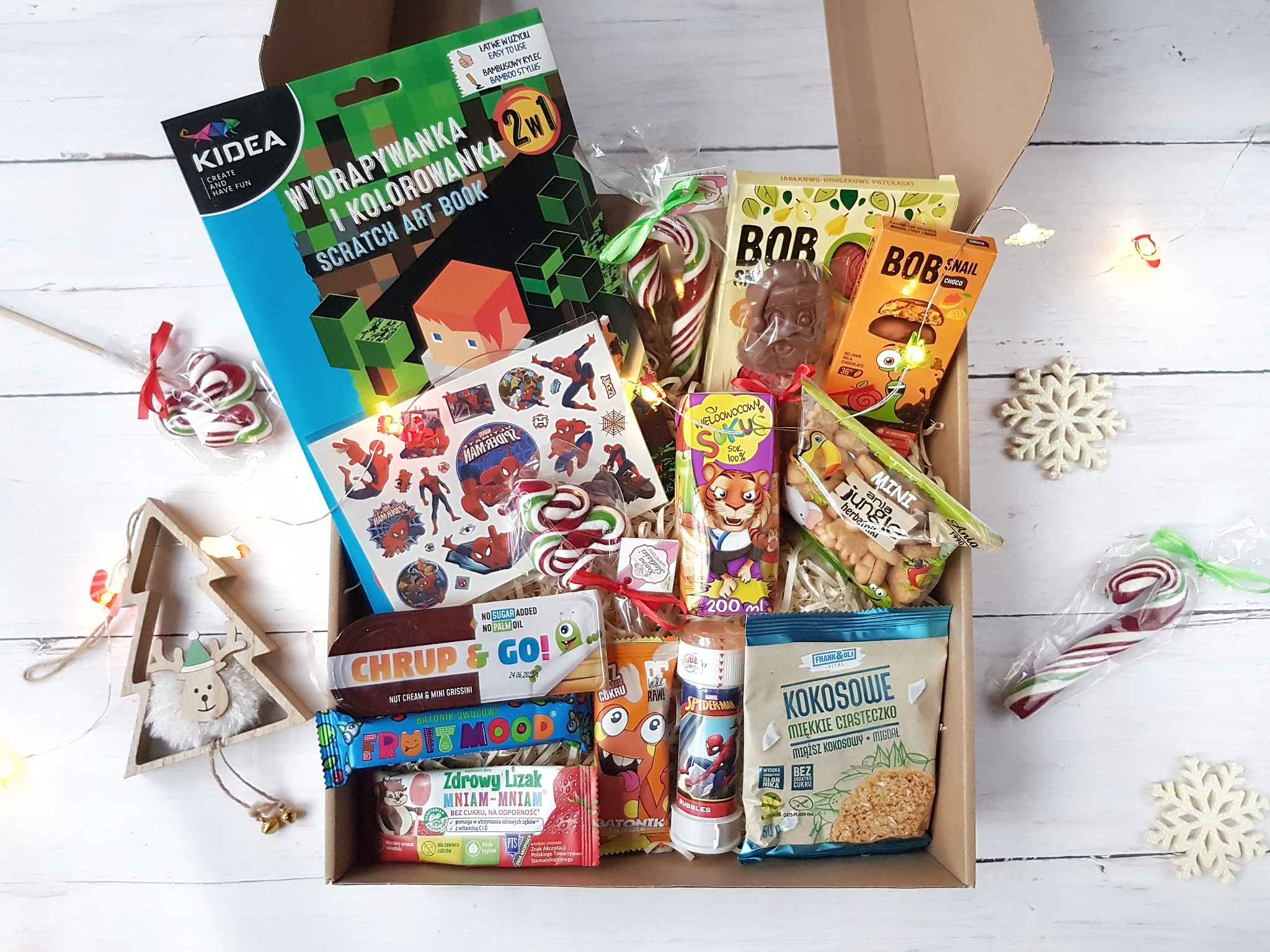 Otwarty karton wypełniony różnorodnymi słodyczami, przekąskami i zabawkami dla dzieci, w tym lizaki, ciasteczka kokosowe, batony owocowe, książeczka do zdrapywania z motywem Minecrafta i naklejki...