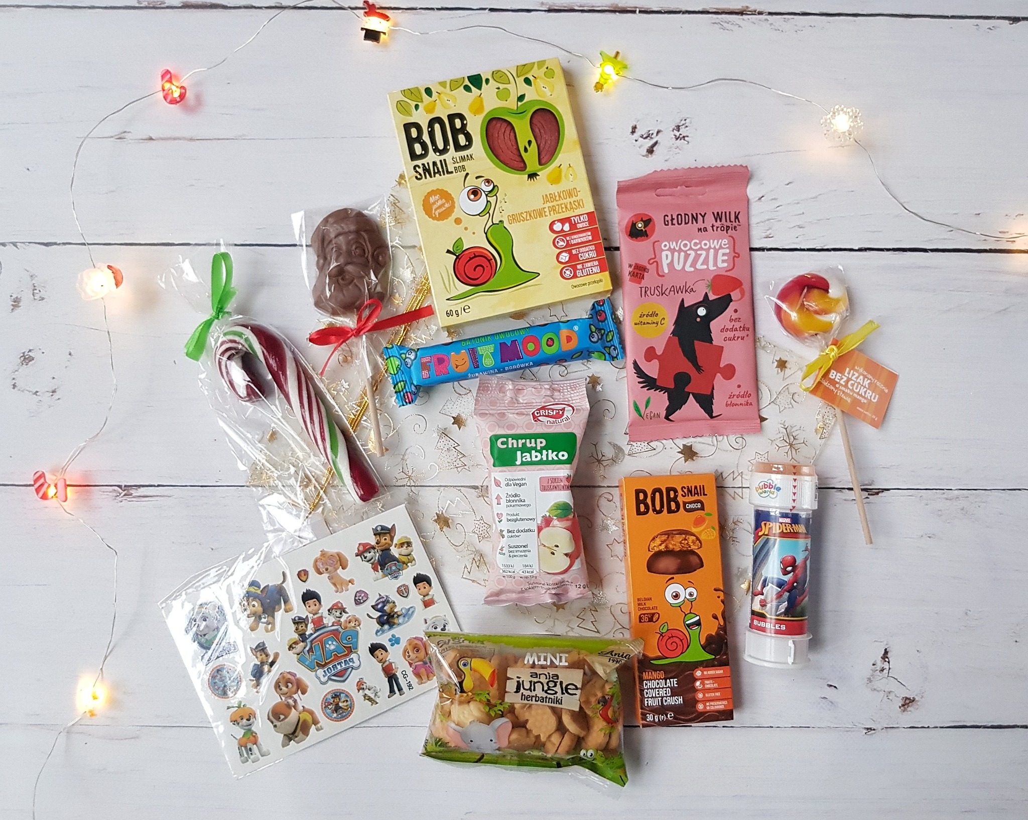 Zestaw słodyczy dla dzieci na białym drewnianym tle, w skład którego wchodzą lizaki, czekoladki, żelki owocowe, naklejki z motywem Psi Patrol oraz bańki mydlane ze Spidermanem, całość oświetlona...