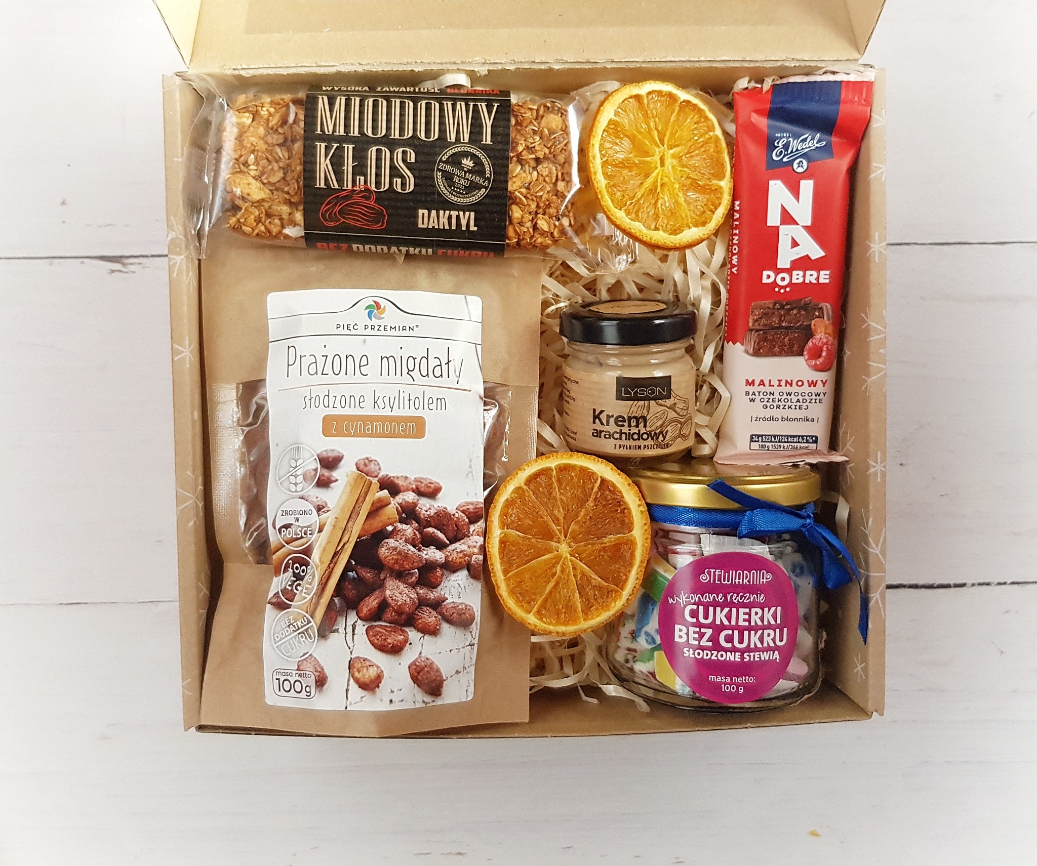 Otwarty kartonowy box wypełniony produktami spożywczymi: baton miodowy, prażone migdały z cynamonem, krem arachidowy z pyłkiem pszczelim, baton malinowy w czekoladzie oraz cukierki bez cukru...