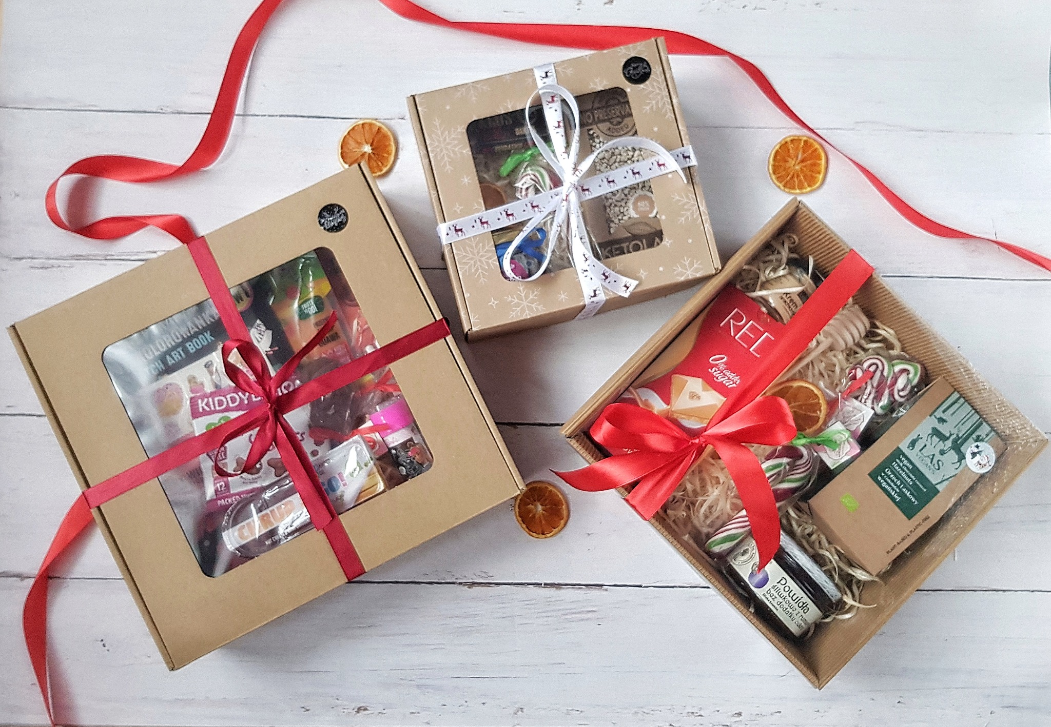 Trzy kosze prezentowe na białym drewnianym tle, każdy z innym motywem: jeden z zabawkami i słodyczami dla dzieci, drugi z produktami spożywczymi z motywem świątecznym, trzeci z produktami...