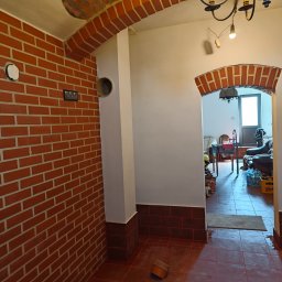 Zbigniew Magdziarz - Wnętrze z surową ceglaną ścianą, otworem wentylacyjnym, i widocznym fragmentem instalacji elektrycznej, z ceglanym łukiem nad przejściem do dalszej części pomieszczenia z widocznymi butelkami wody...