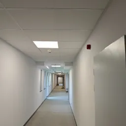 Długi, pusty korytarz z białymi ścianami, panelowym sufitem z wbudowanym oświetleniem i betonową podłogą, widok w perspektywie z alarmem przeciwpożarowym na ścianie.