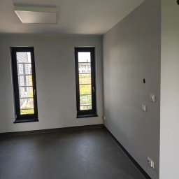 Zbigniew Magdziarz - Pomalowane na szaro puste pomieszczenie z dwoma pionowymi oknami w czarnych ramach, widocznymi gniazdkami i włącznikami na ścianie oraz ciemną podłogą.