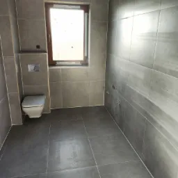 Szara łazienka w trakcie wykańczania: widoczne płytki na ścianach i podłodze, częściowo zamontowana toaleta wisząca, okno z drewnianą ramą oraz przygotowane punkty instalacyjne na ścianie.