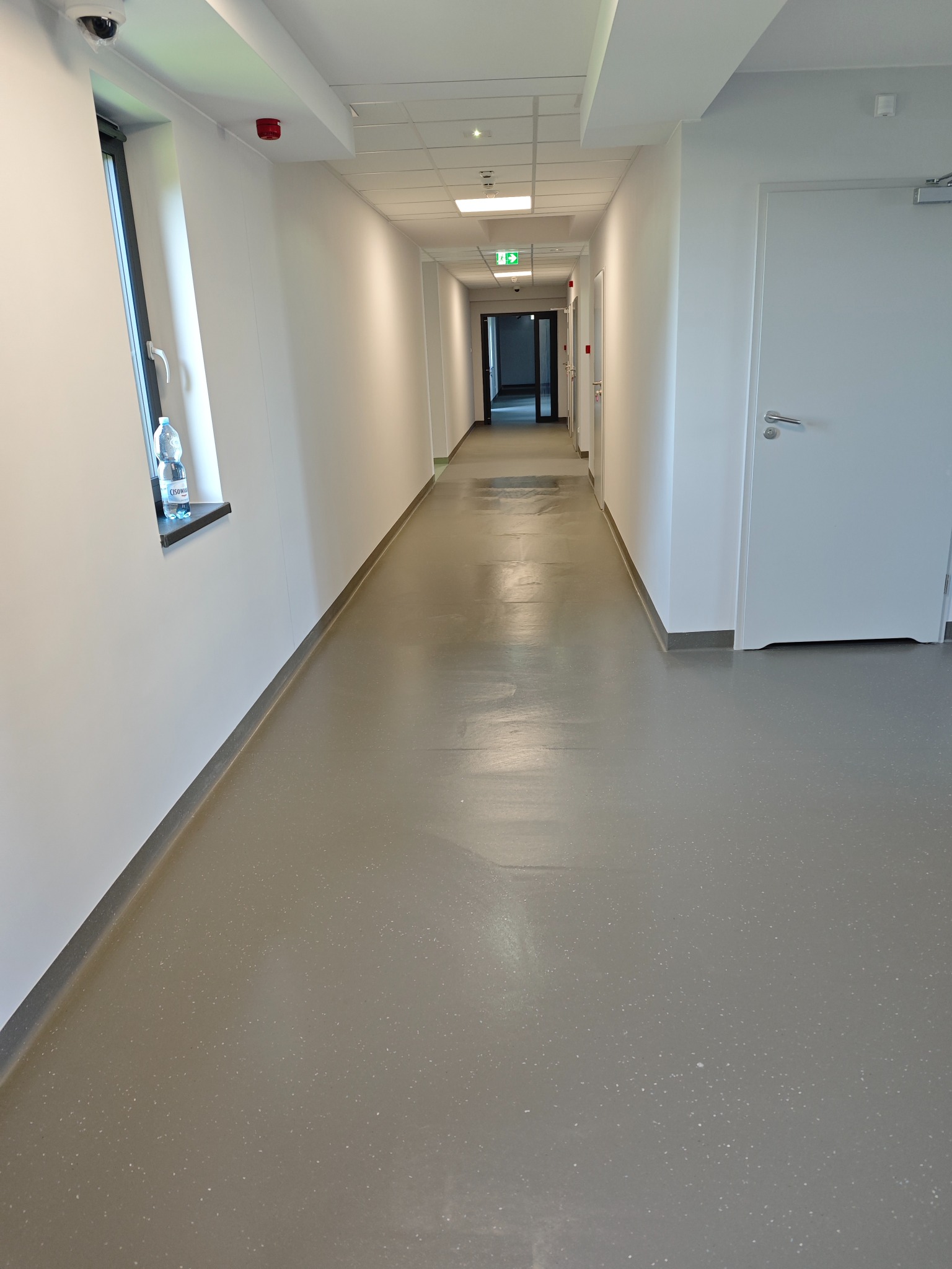 Długi, jasny korytarz w budynku użyteczności publicznej z szarą podłogą, białymi ścianami, oknem z butelką wody, kamerą przemysłową i drzwiami przeciwpożarowymi.