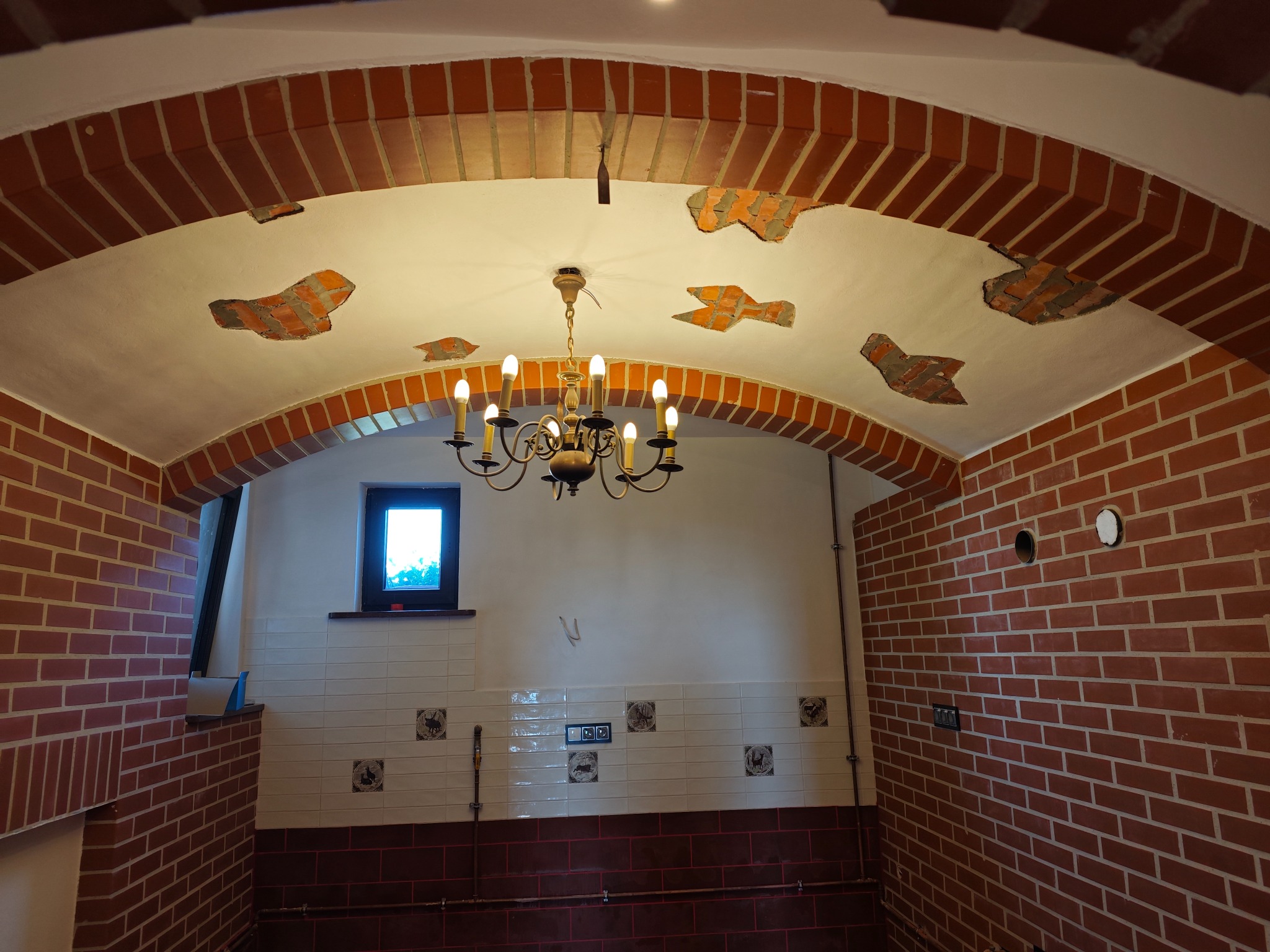 Wnętrze pomieszczenia z ceglanym sklepieniem łukowym, białymi ścianami z dekoracyjnymi płytkami i wiszącym, klasycznym żyrandolem.