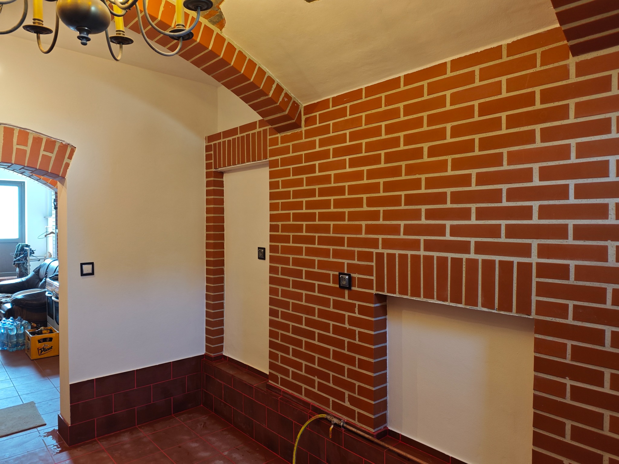 Wnętrze z ceglaną ścianą, łukami i obniżonym ceglanym cokołem; widoczna instalacja wodna z zaworem.