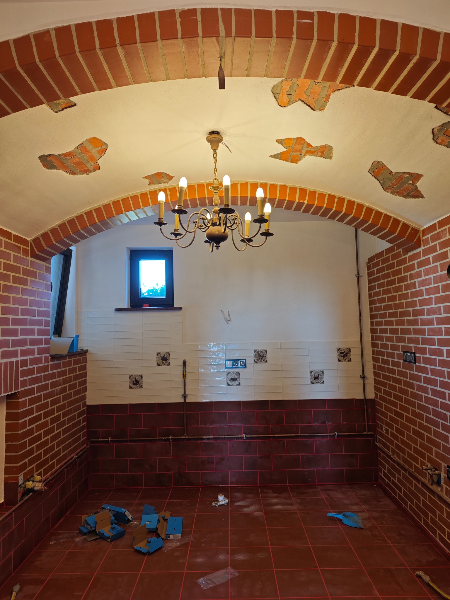Wnętrze pomieszczenia z łukowym sufitem wykończonym cegłą, fragmentami odsłoniętej cegły na suficie, ceramicznymi płytkami na ścianach i podłodze, oraz wiszącym żyrandolem w trakcie prac...