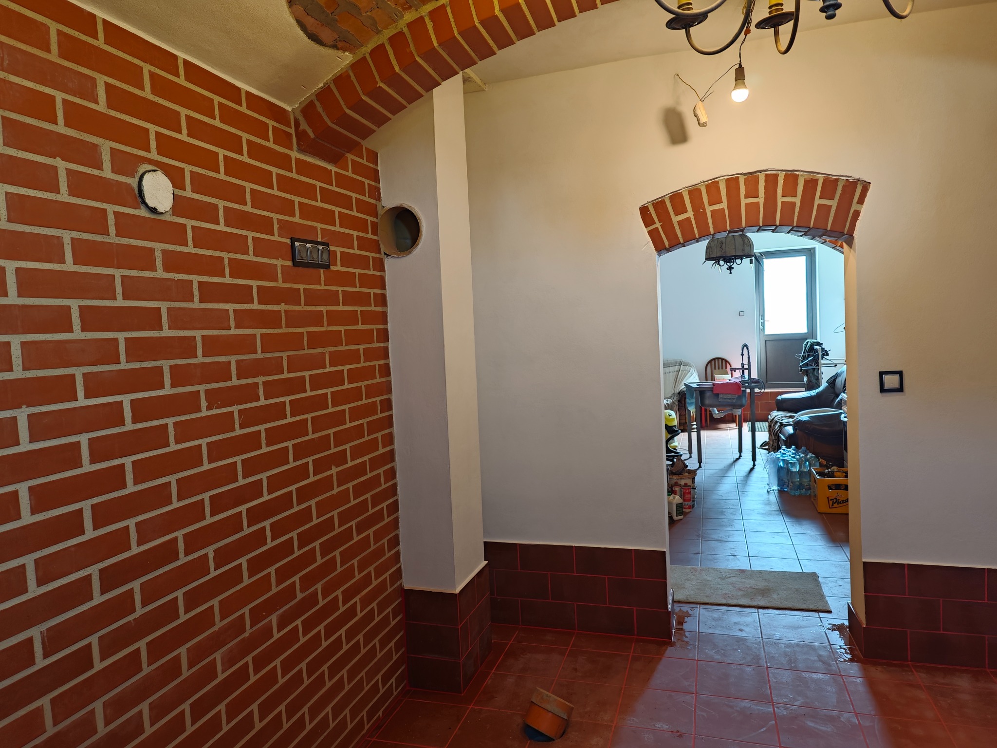 Wnętrze z surową ceglaną ścianą, otworem wentylacyjnym, i widocznym fragmentem instalacji elektrycznej, z ceglanym łukiem nad przejściem do dalszej części pomieszczenia z widocznymi butelkami wody...