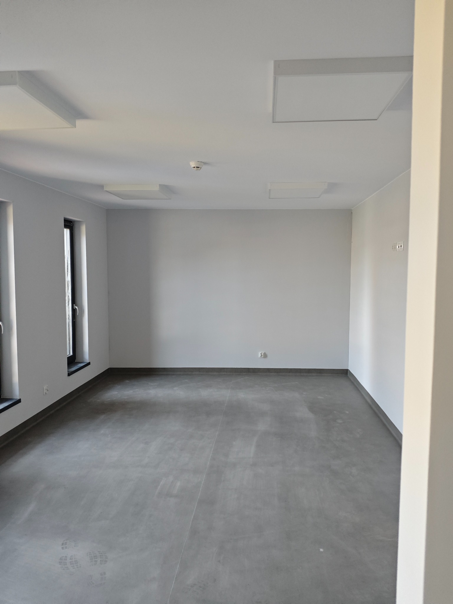 Puste, szare pomieszczenie z betonową podłogą, trzema oknami z czarnymi ramami, białymi ścianami i kwadratowymi panelami oświetleniowymi na suficie. Na podłodze widoczne ślady butów.