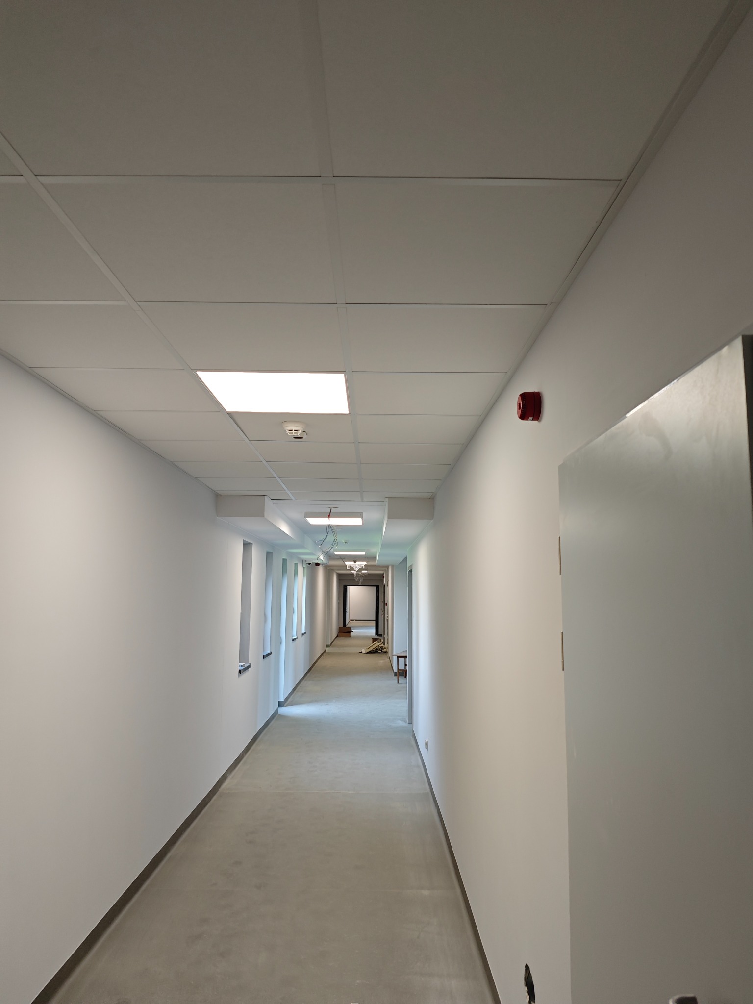 Długi, pusty korytarz z białymi ścianami, panelowym sufitem z wbudowanym oświetleniem i betonową podłogą, widok w perspektywie z alarmem przeciwpożarowym na ścianie.