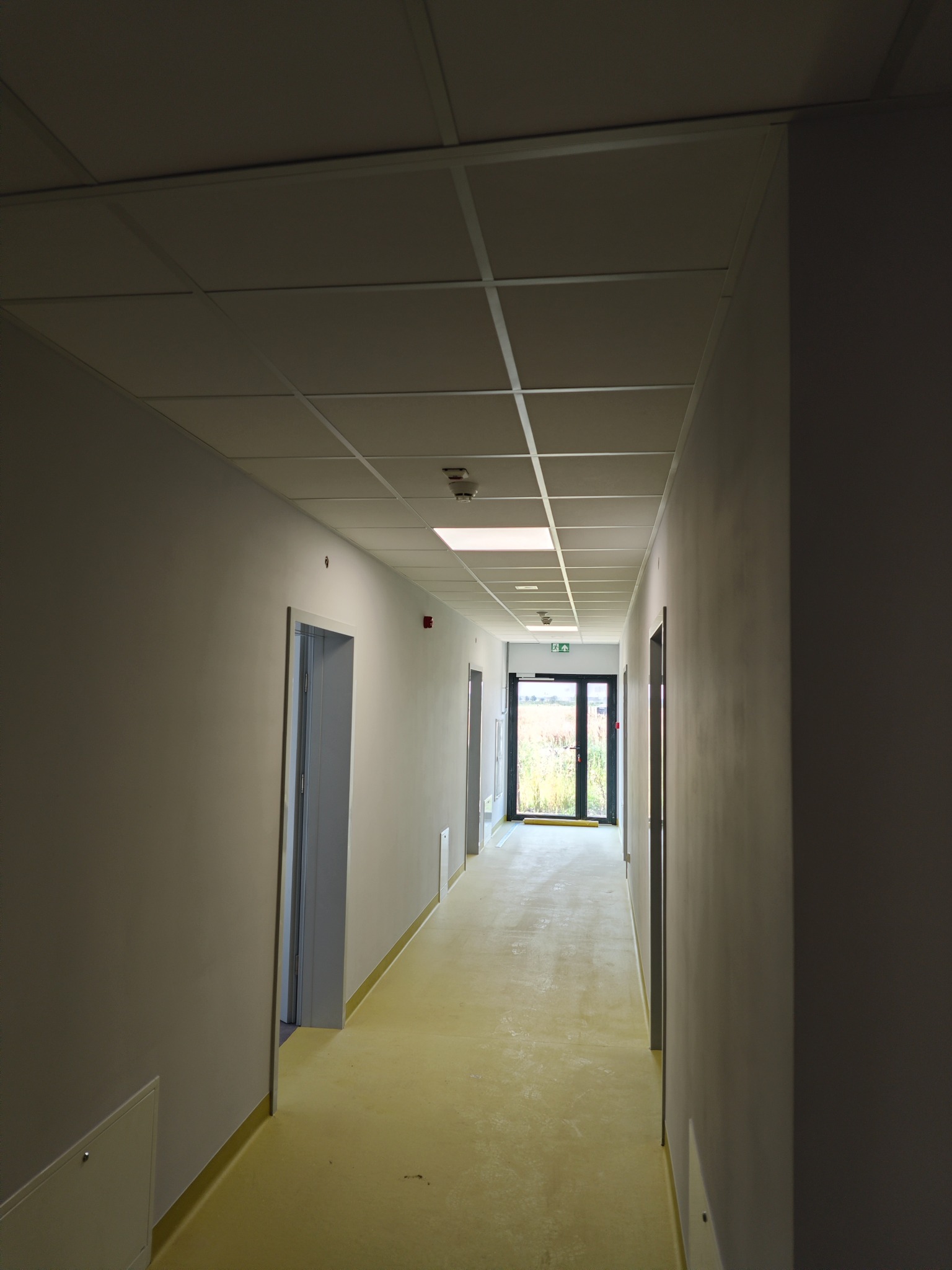 Długi korytarz z panelami sufitowymi i żółtą podłogą, widoczne drzwi ewakuacyjne na końcu, ściany pomalowane na szaro.