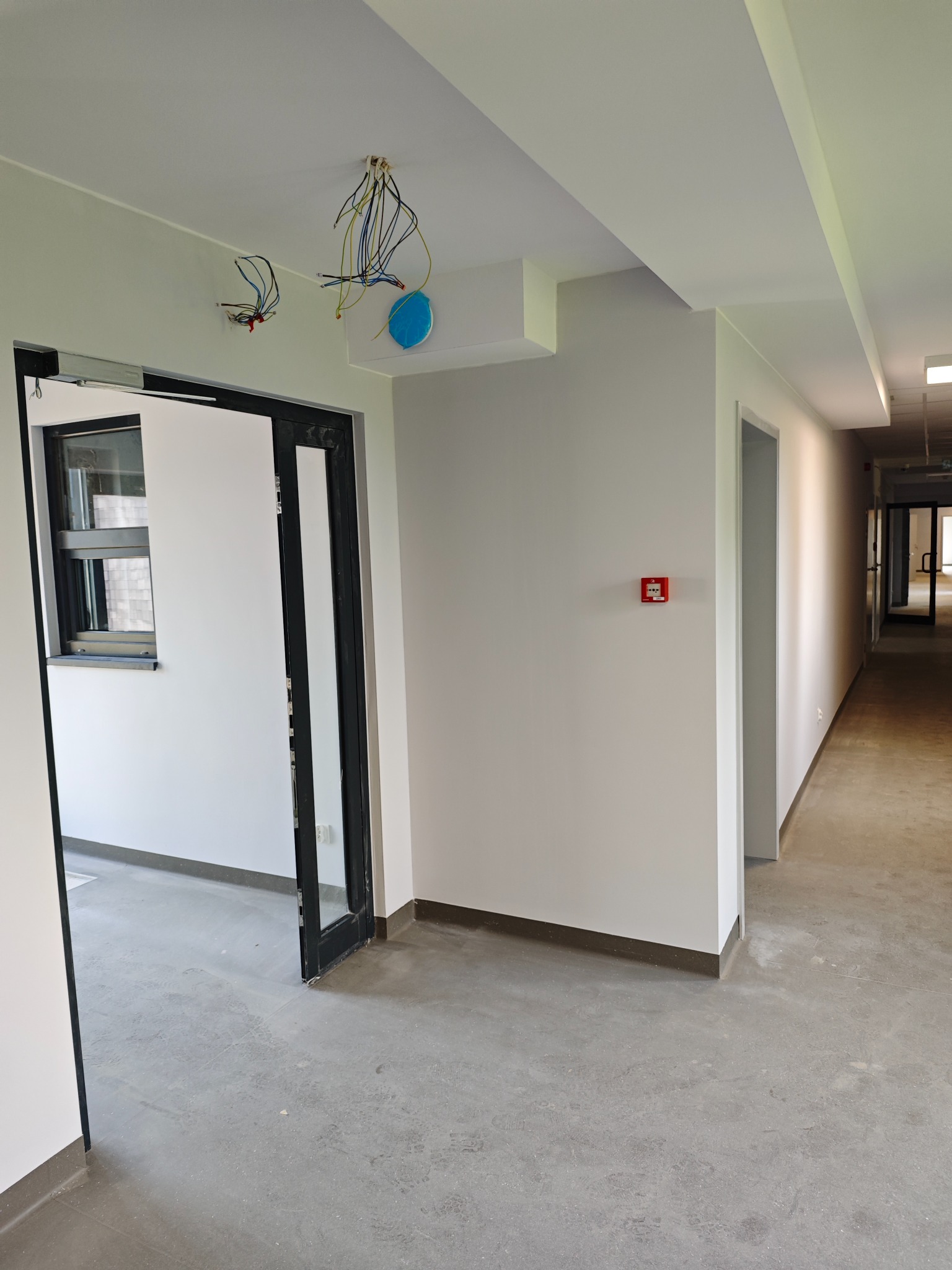 Wnętrze w trakcie prac: widoczne kable wystające z sufitu, świeżo pomalowane ściany na biało, surowa podłoga, otwarte drzwi z czarną ramą i małym oknem, oraz czujnik dymu na ścianie w korytarzu.