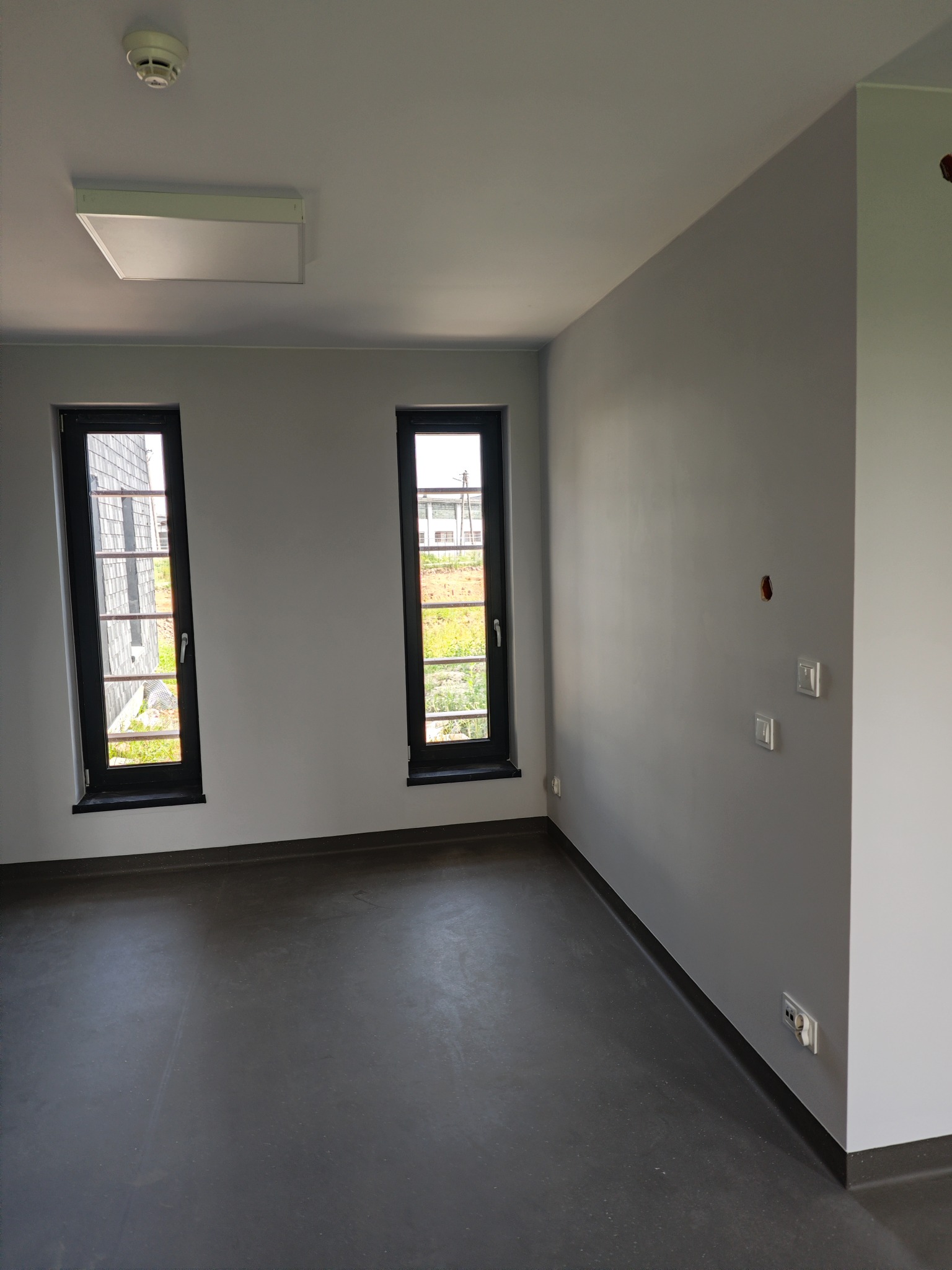 Pomalowane na szaro puste pomieszczenie z dwoma pionowymi oknami w czarnych ramach, widocznymi gniazdkami i włącznikami na ścianie oraz ciemną podłogą.
