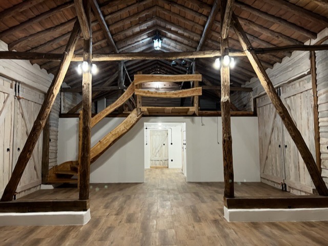 Przestronne wnętrze stodoły zaadaptowanej na salę do ćwiczeń, z drewnianą konstrukcją stropu, schodami na antresolę i podłogą imitującą drewno.