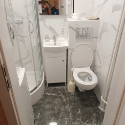 REM-RYS - Nowoczesna, mała łazienka z kabiną prysznicową, umywalką z szafką, toaletą i marmurowymi płytkami na ścianach i podłodze. W lustrze odbija się osoba robiąca zdjęcie oraz starsza kobieta.