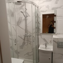 REM-RYS - Nowoczesna łazienka z kabiną prysznicową typu walk-in z chromowanymi elementami, umywalką z szafką w kolorze białym oraz płytkami imitującymi marmur na ścianach; w lustrze odbija się osoba robiąca...