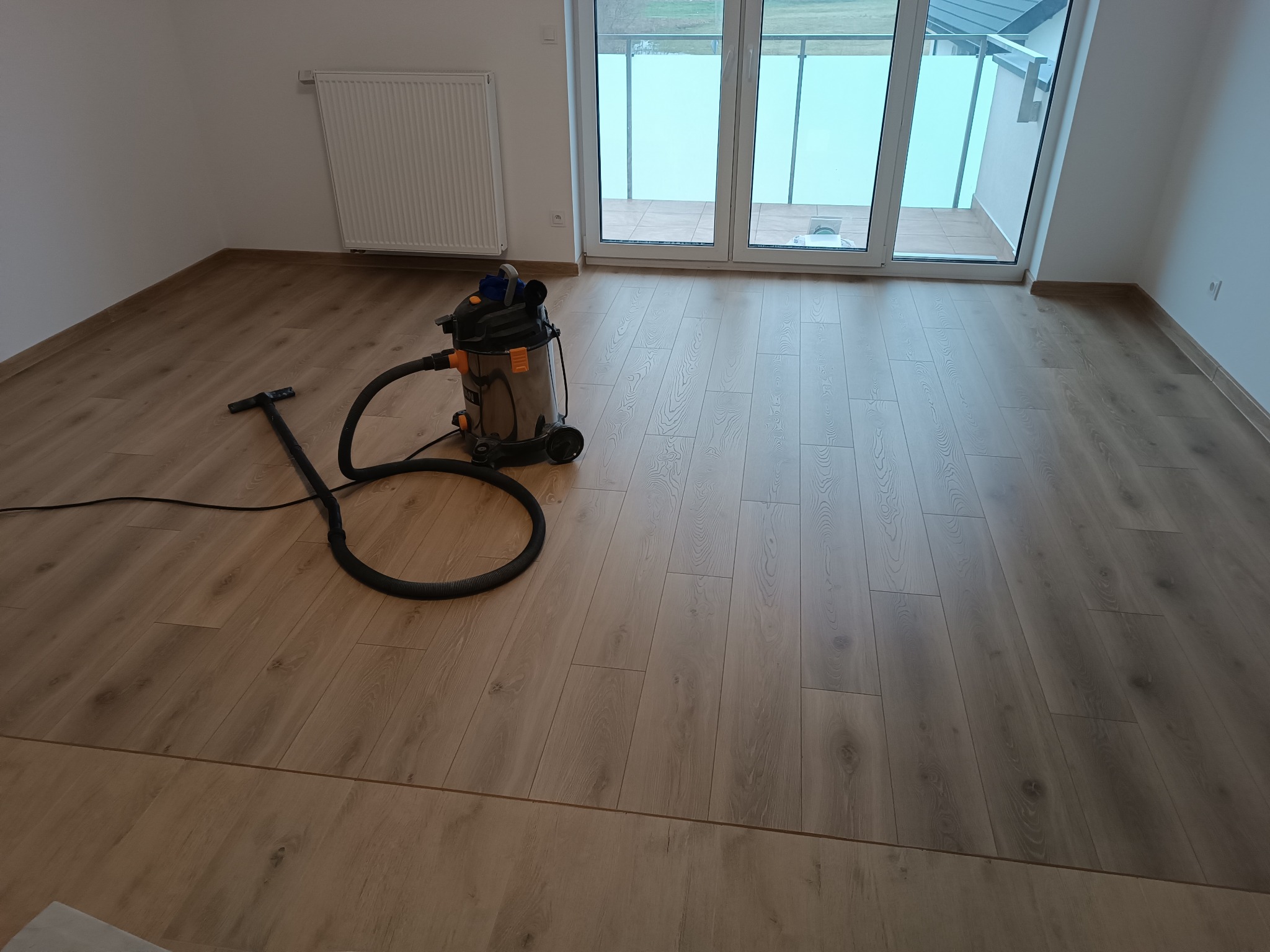 Świeżo ułożone panele podłogowe w jasnym pokoju, z widocznym odkurzaczem przemysłowym i wyjściem na balkon.