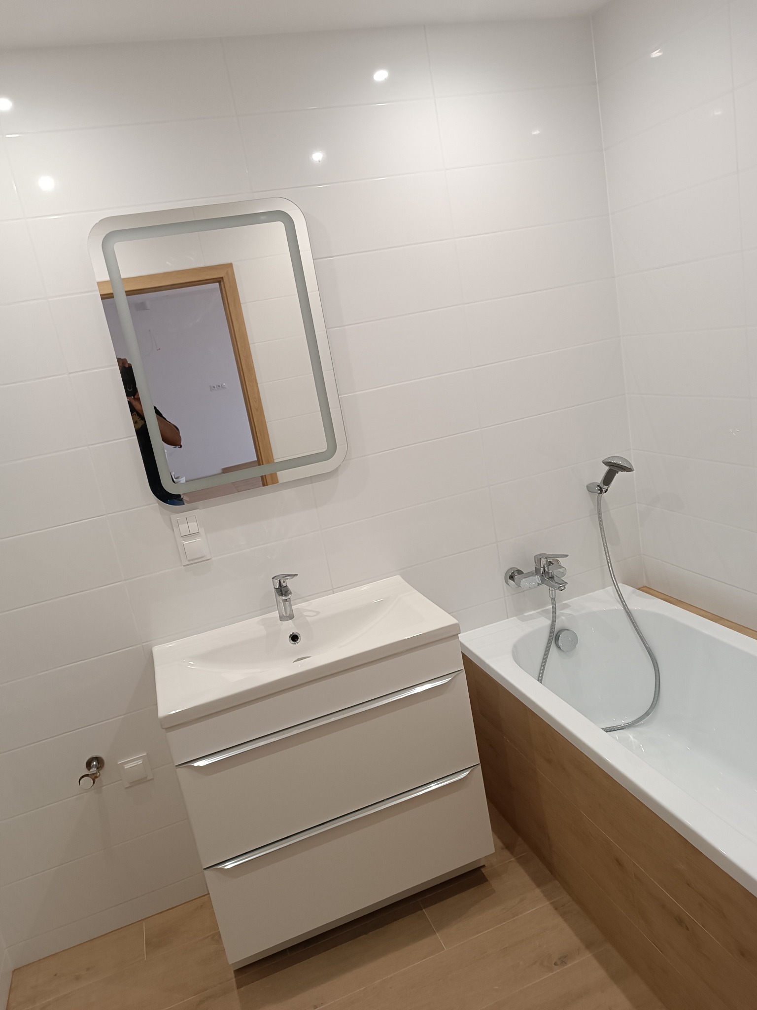 Nowoczesna łazienka z białymi płytkami, podświetlanym lustrem, białą szafką pod umywalką i wanną z drewnopodobną obudową.