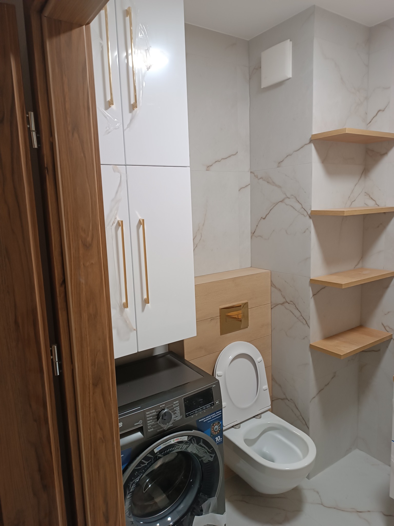 Wykończona łazienka w Białymstoku z marmurowymi płytkami, drewnianymi półkami, wiszącą toaletą z deską oraz pralką pod szafką z złotymi uchwytami, jeszcze w folii ochronnej.