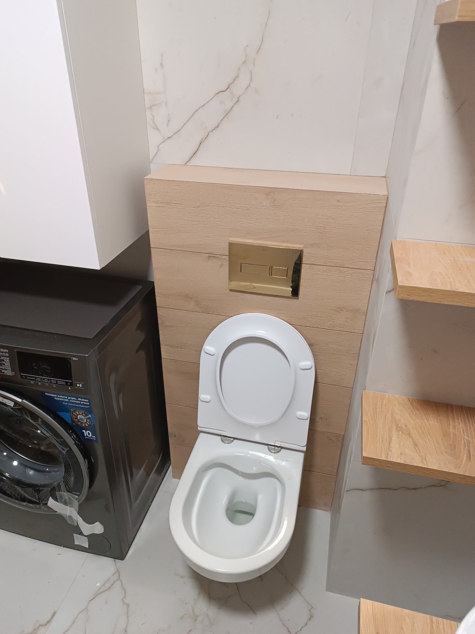 Nowoczesna toaleta wisząca z drewnopodobną obudową stelaża i złotym przyciskiem spłukującym, sąsiadująca z pralką i marmurową ścianą w łazience.