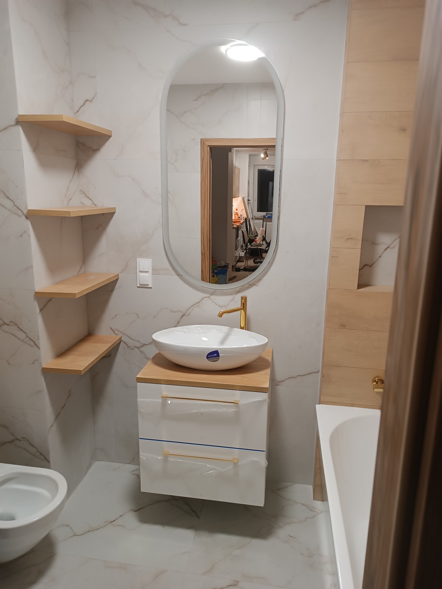Nowoczesna łazienka z umywalką nablatową na szafce, owalnym lustrem, drewnianymi półkami i marmurowymi płytkami na ścianach i podłodze. Elementy wykończeniowe w kolorze złotym.