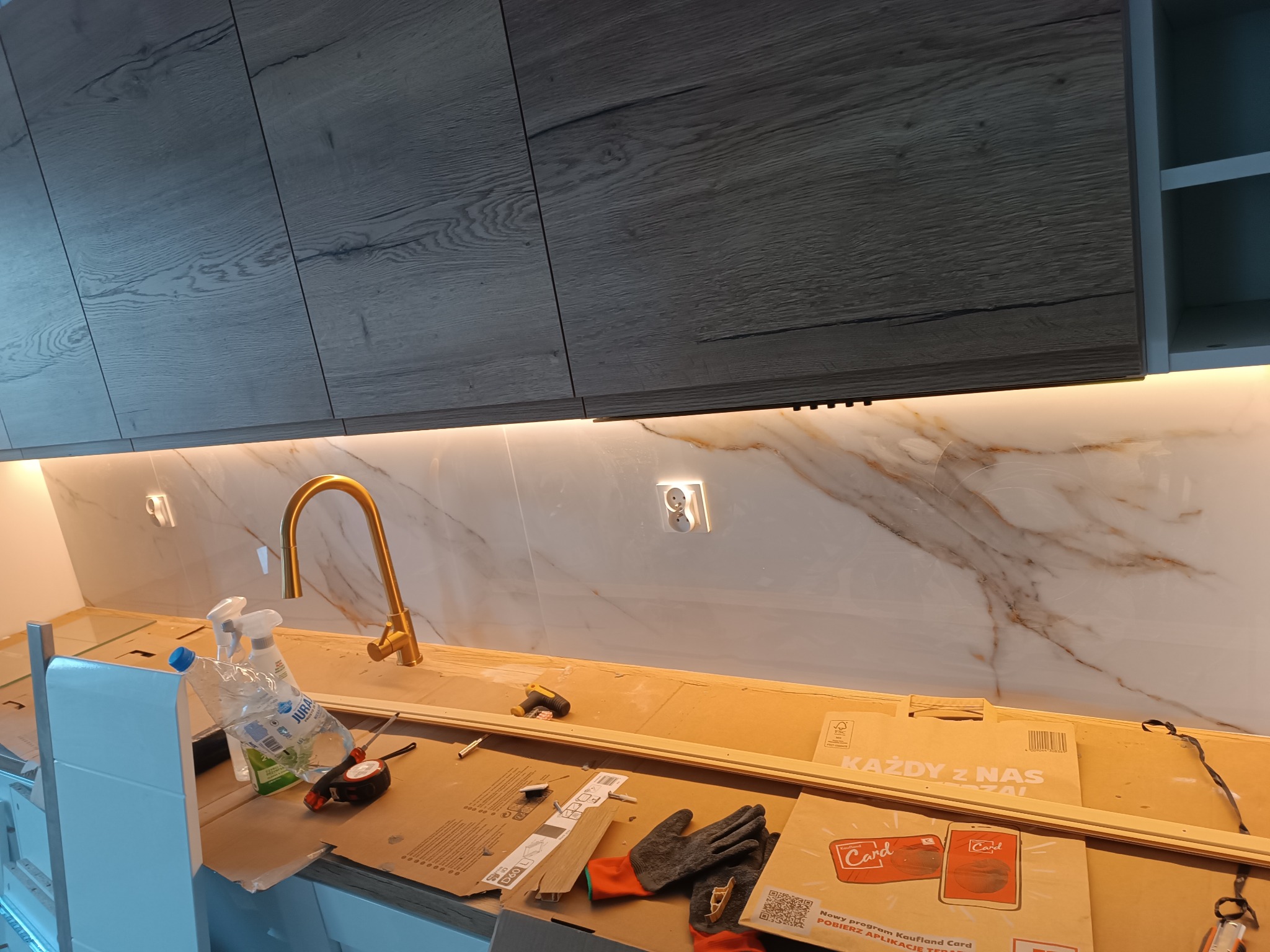 Montaż złotej baterii kuchennej na tle marmurowej ściany, widoczne narzędzia, kartony i rękawice robocze na blacie.