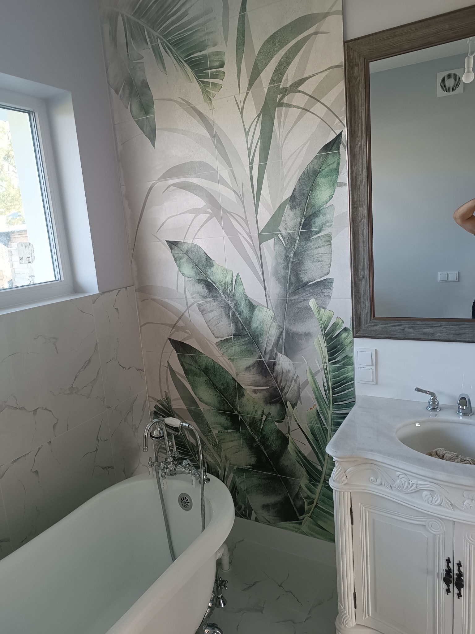 Elegancka łazienka z wolnostojącą wanną na nóżkach, marmurowymi płytkami i szafką umywalkową w stylu retro, wyróżniająca się tapetą z motywem tropikalnych liści.